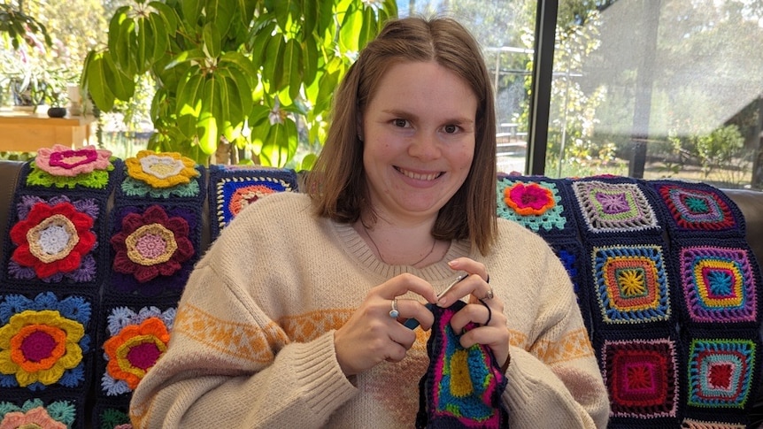 A young woman sits on a couch with a crochet square blanket on it, smiling and crocheting