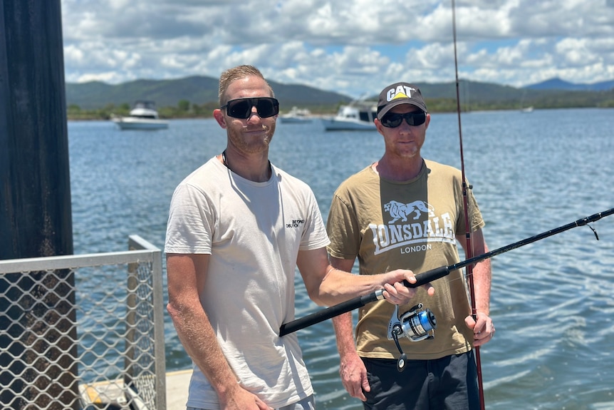 Two men with fishing rods smile at the camera in front of water