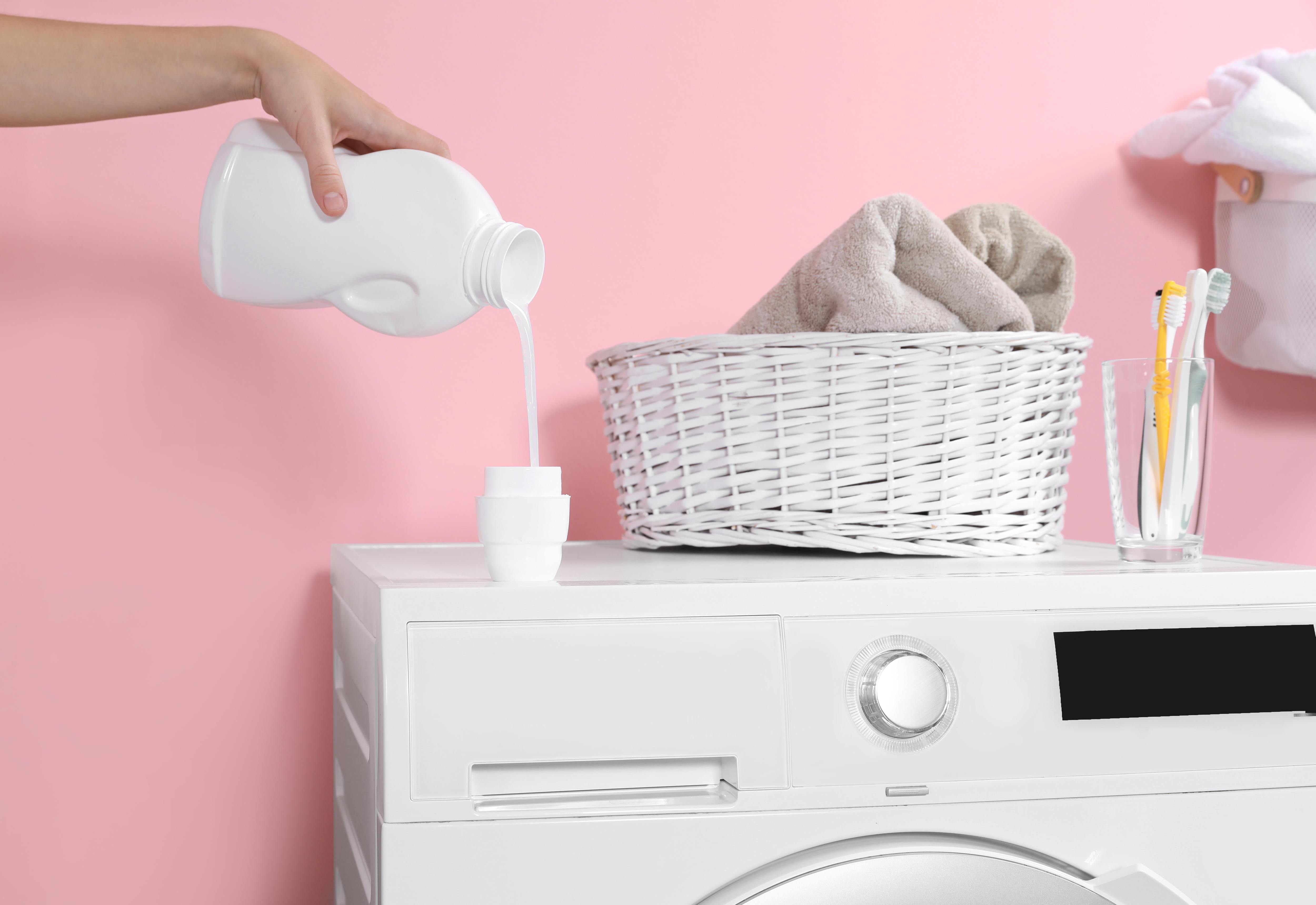 A woman pours laundry liquid into its lid on top of a washing machine, against a pink background.