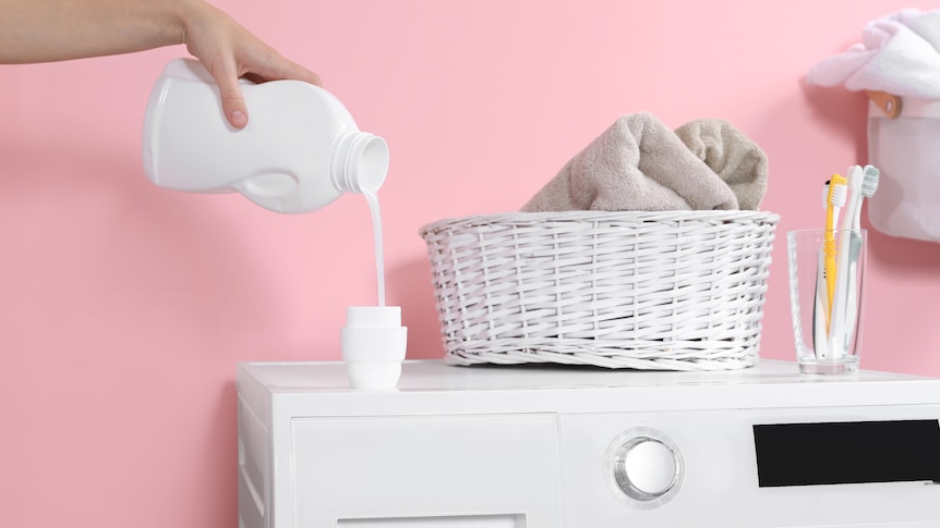 A woman pours laundry liquid into its lid on top of a washing machine, against a pink background.