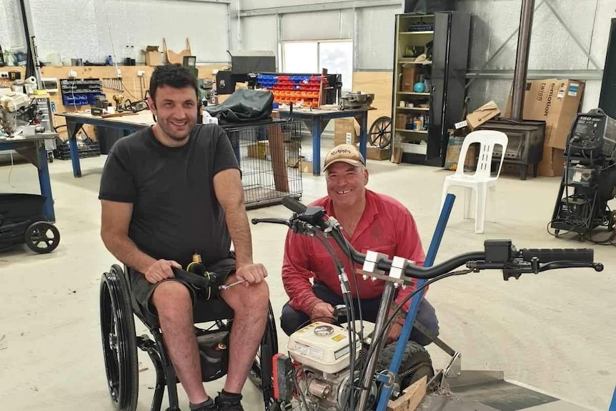 Two smiling men – one in a wheelchair – in a large shed.