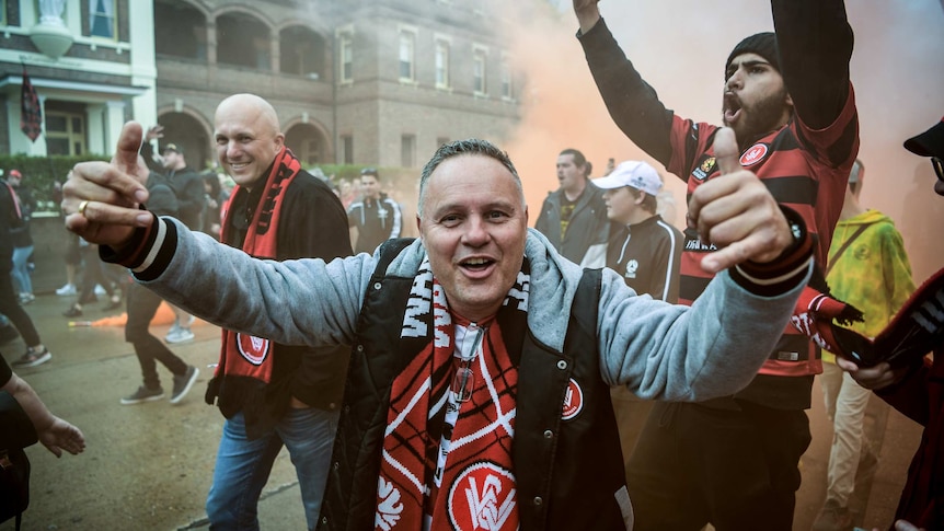 A man smiles into the camera as flares go off in the background.