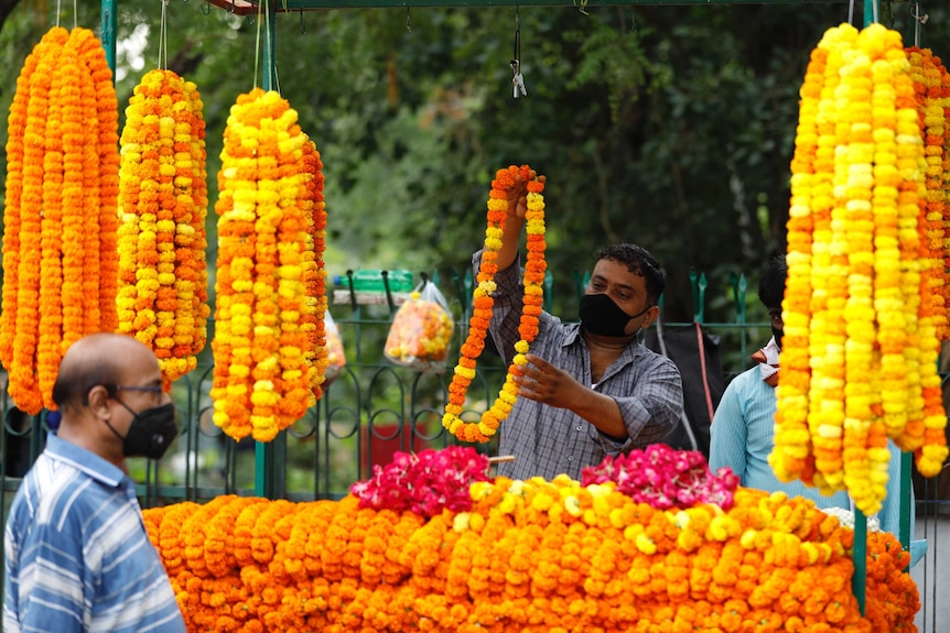 A man wearing a face mask arranges loops of yellow and orange flowers at a roadside stall outside a temple as people walk past.