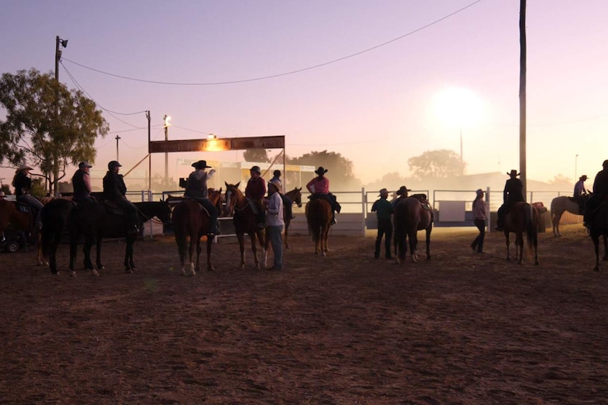 Riders on horseback wait in front of a dusty arena