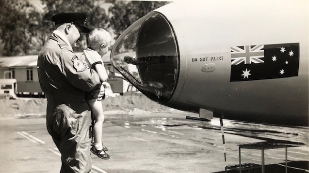 black and white photo of a man in Airforce overalls lifting a young child to look at a plane