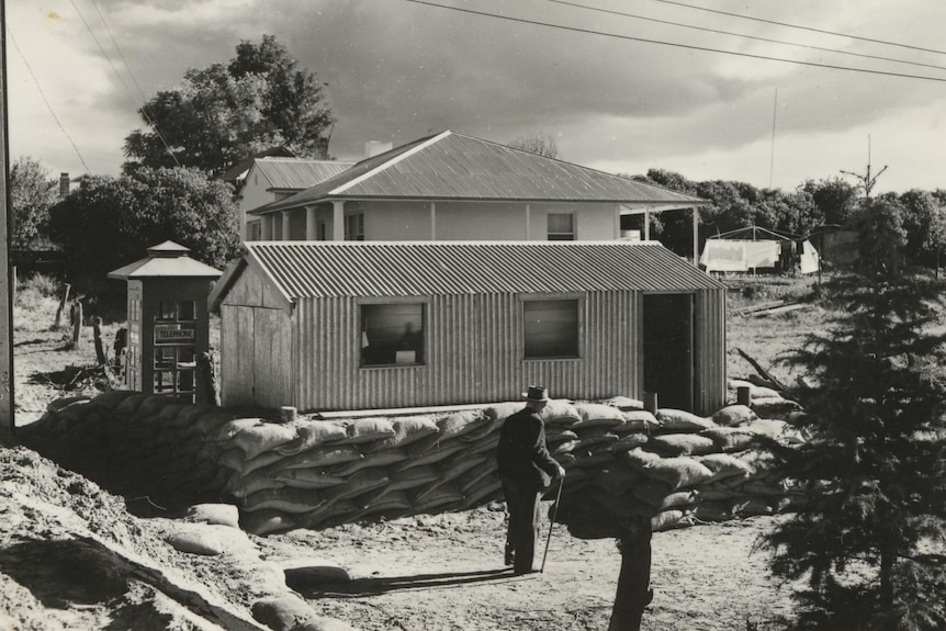 A black and white phot, shows a tin shed surrounded by sanbags, a man with walking stick watches on