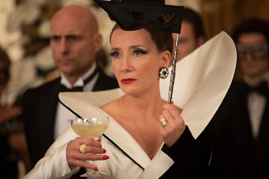 Film still of Emma Thompson as The Baroness wearing a structured dress, holding a martini glass and eye mask in Cruella