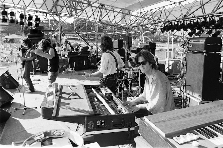 Black and white: Band on stage playing in front of outdoor crowd. Cold Chisel keyboard player Don Walker wearing sunglasses