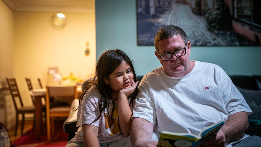 A man in glasses and a white t-shirt holds a book out as a younger girl reads aloud and leans into him in their living room.