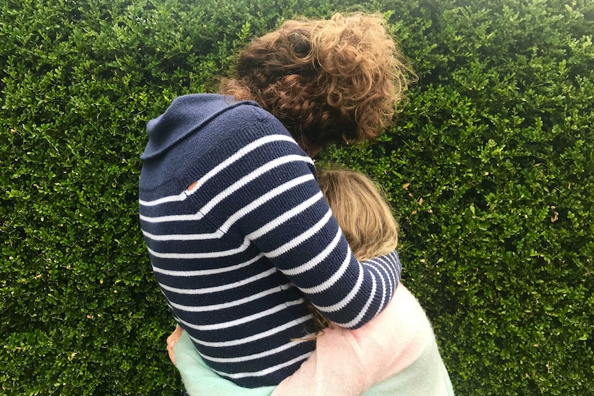 The back view of a woman hugging a child in front of a hedge.