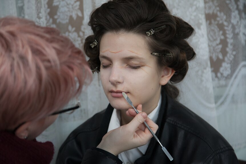 A make-up artist applies make-up to a young woman's face using a thin brush..