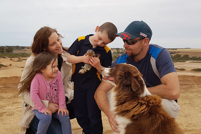 A mother, father, daughter and a son holding a lizard stand on a dirt hill with a brown and white long-haired dog.