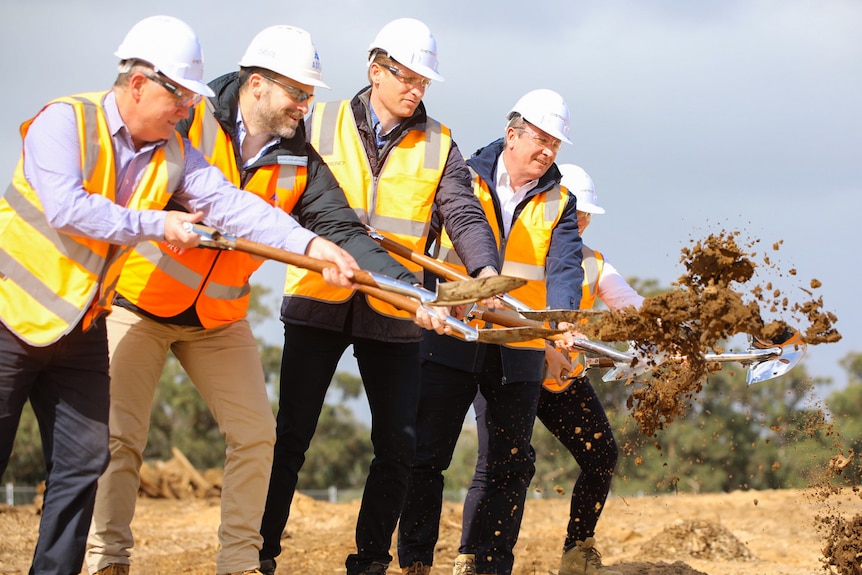 Four men in high-vis vests and hard hats throwing shovels of dirt towards the right of frame.