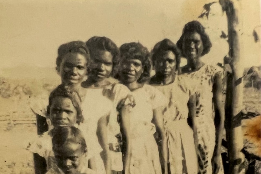 A row of women stand side by side at a cattle station. They are all wearing cotton dresses and are happy.