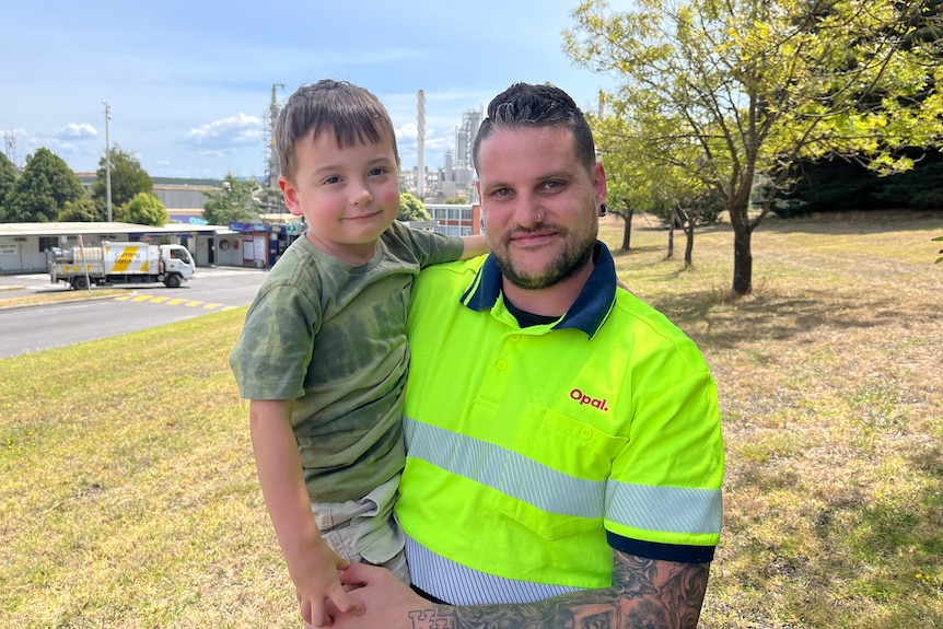 A man wearing a hi-vis shirt, standing outside holding his young son.