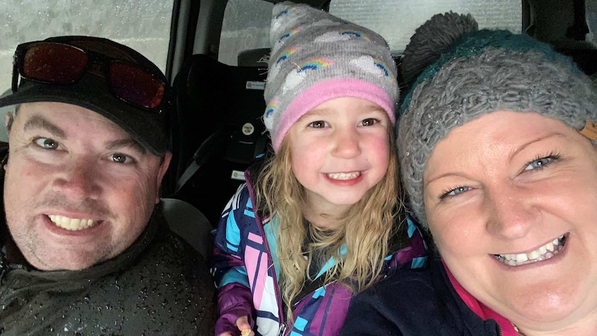 A man and woman sitting in a car with their young daughter between them, wearing a beanie, all smiling.