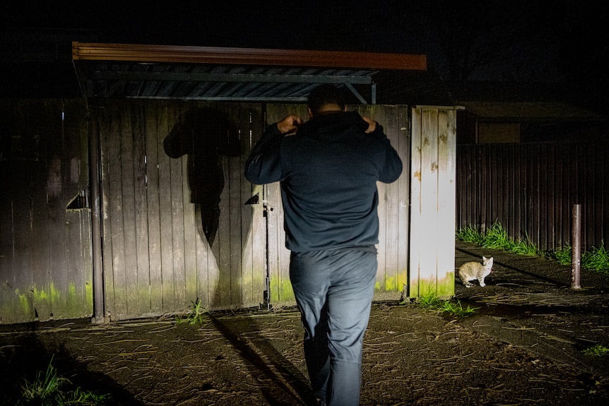 A man walks at night. A car headlight shines his shadow at a fence.
