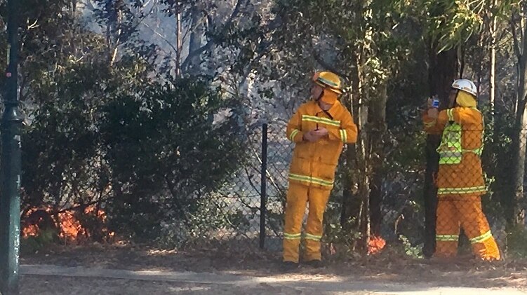 Two firefighters stand in scrub waith fire at their feet.