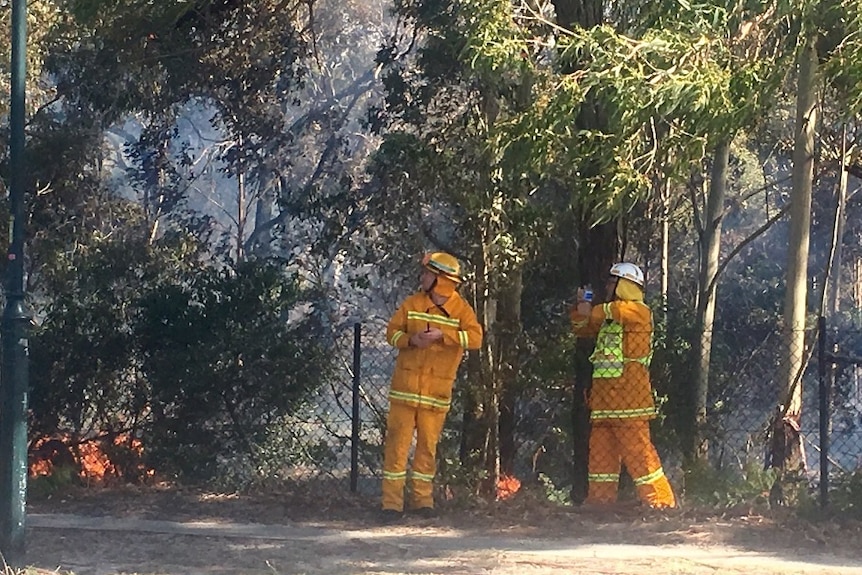 Two firefighters stand in scrub waith fire at their feet.