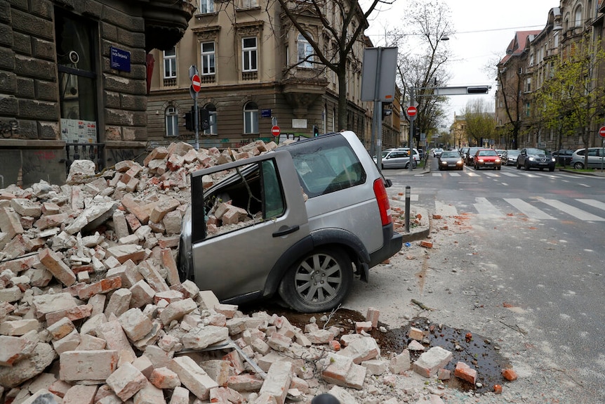 Un SUV plateado destrozado se ve debajo de una pila de ladrillos en una calle antigua rodeada de patrimonio en un día nublado.