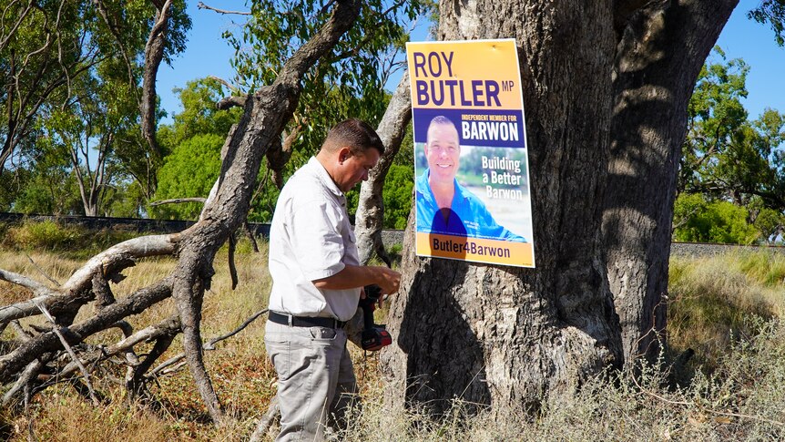 A man stands among tall grass as he nails a political sign to a tree