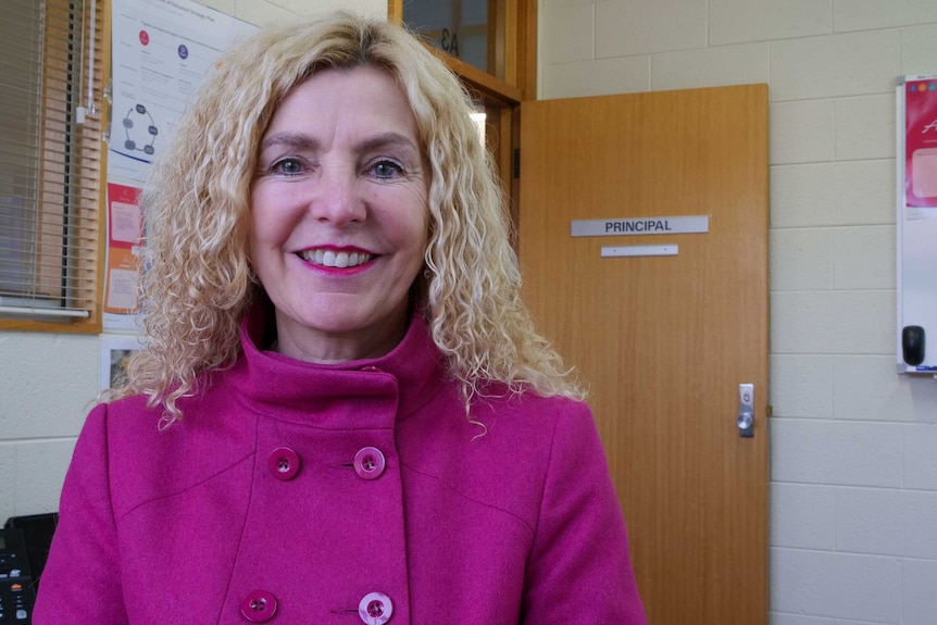 A woman wearing a pink jacket smiling at the camera, a door behind her says 'principal'