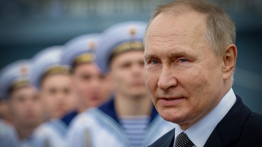 Vladimir Putin with a slight smile on his face, with a row of men in naval uniforms lined up behind him 