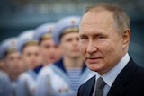 Vladimir Putin with a slight smile on his face, with a row of men in naval uniforms lined up behind him 