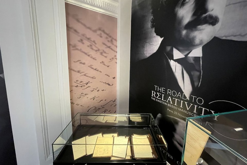 전경에 있는 박물관의 유리 케이스에 새겨진 문서, 배경에 아인슈타인의 상대성 이론의 포스터