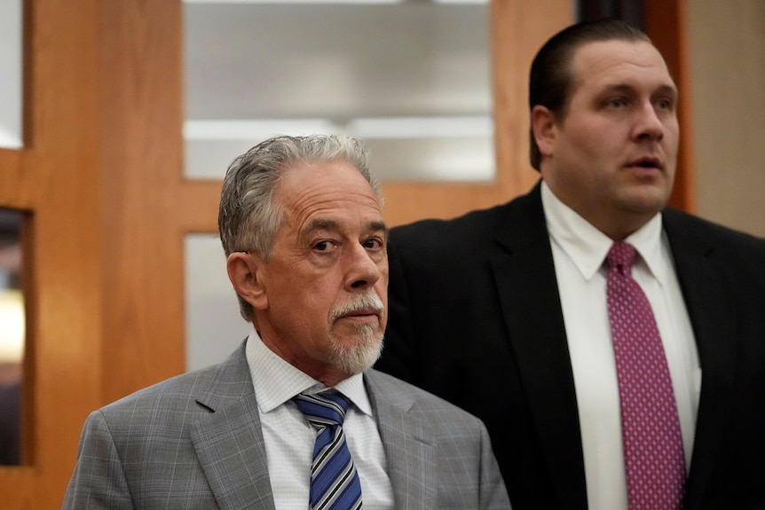 A man with grey hair and a white goatee is pictured wearing a grey suit. Behind him a larger man wearing a black suit. 