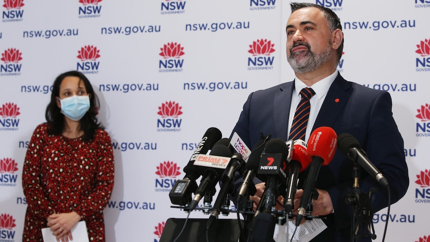 A bearded politician in a dark suit – John Barilaro – speaking to the media, flanked by the Deputy CHO, who is wearing a mask.