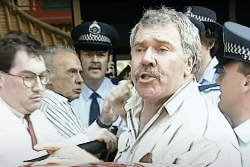 Screengrab of a grey-haired man being wrestled into a car by a few policeman 