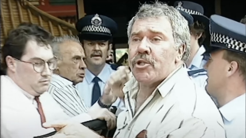 Screengrab of a grey-haired man being wrestled into a car by a few policeman 