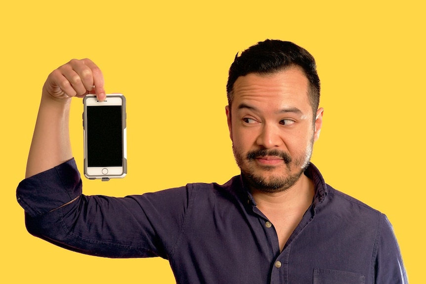 Photo of ABC journalist Jason Om holding up a phone on a yellow background. He's faced racism on gay dating apps like Grindr.
