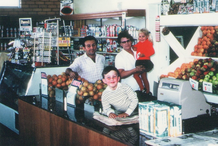 A colour photo from the late 1960s of two parents with a young boy and girl in a store with fruit and cash register.