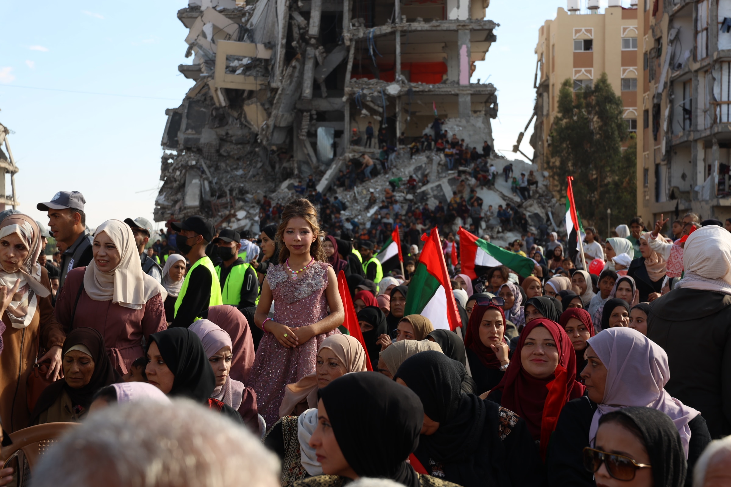 A young girl wearing a dress and makeup stands on a chair amongst a crowd of people in front of a collapsed building.