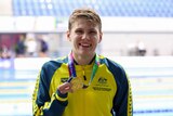 A smiling Australian swimmer stands on the pooldeck with a gold medal around his neck after his event.