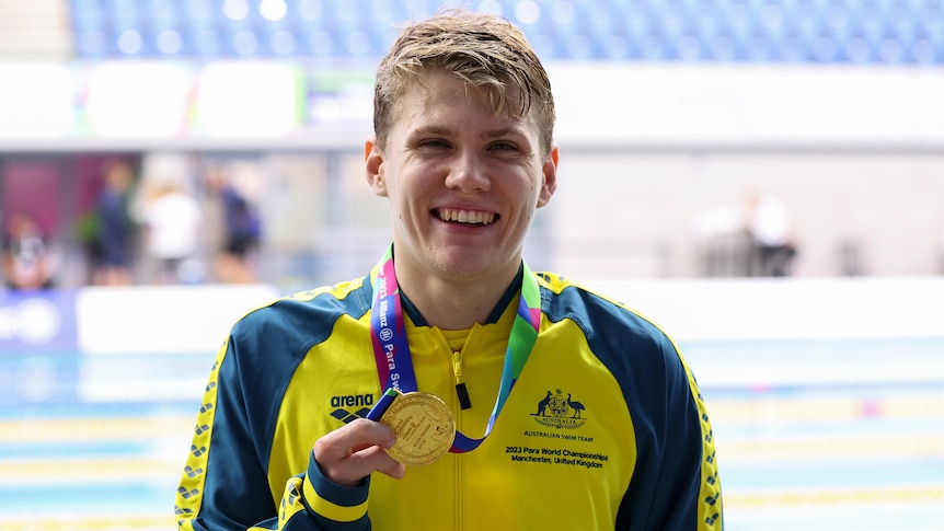 A smiling Australian swimmer stands on the pooldeck with a gold medal around his neck after his event.