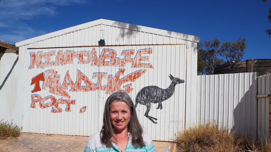 A lady stands in front of a shed with a painted kangaroo and a sign which reads 'Mintabie Trading Post'.