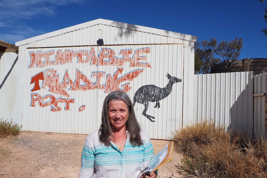 A lady stands in front of a shed with a painted kangaroo and a sign which reads 'Mintabie Trading Post'.