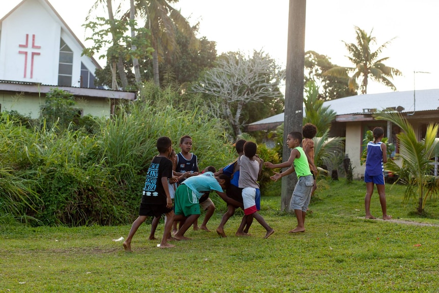 A group of boys play rugby on grass in front of a church with palm trees.