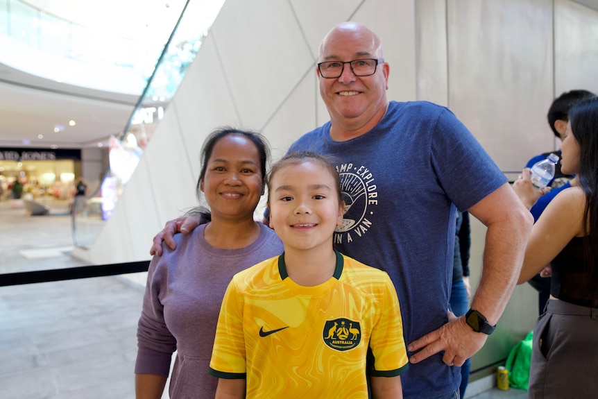 A mum and a dad with their daughter who is wearing a yellow Australian football shirt