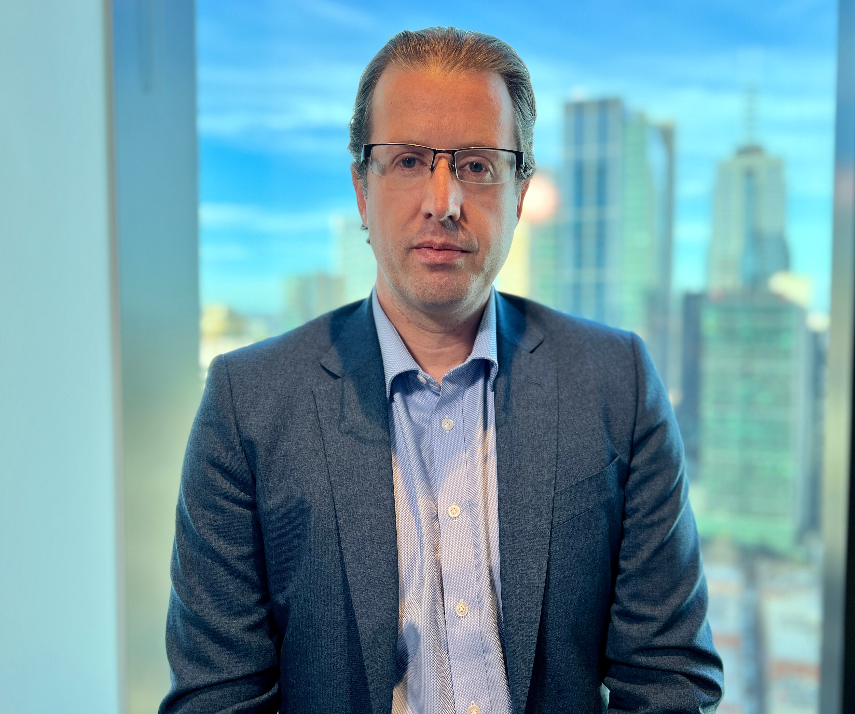 A man wearing a blazer and blue shirt and glasses stands in front of a window with skyscrapers behind him on a sunny day.