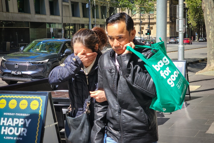 Mr Vo wears a black leather jacket and carries a green shopping bag while his wife holds his arm and covers her face.