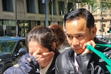 Mr Vo wears a black leather jacket and carries a green shopping bag while his wife holds his arm and covers her face.