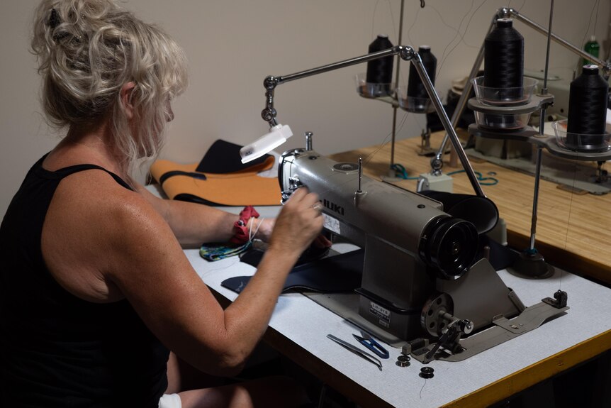 A grey-haired woman with a tousled bun sits and sews on a machine 