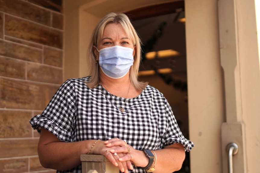 Blonde woman in gingham shirt wears a mask and stands in the doorway of her store.