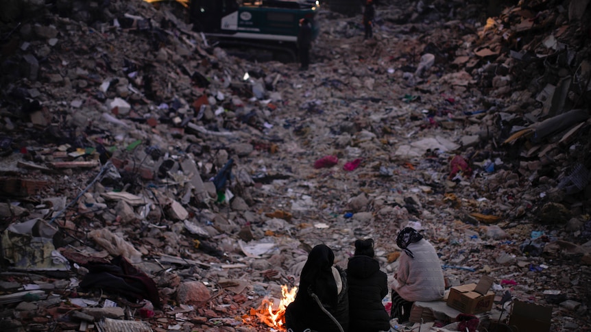 Three people sit among the rubble in from of a fire. 