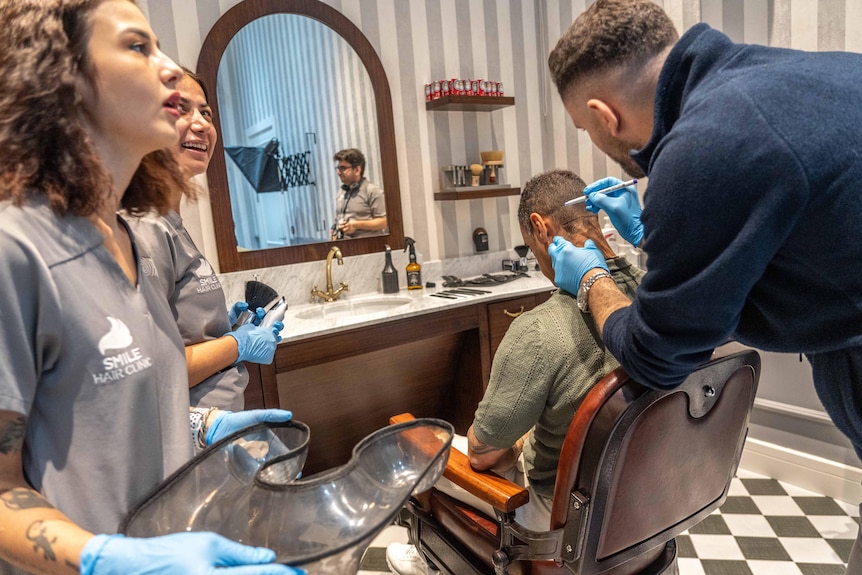 A man sits in a barber chair while someone wearing surgical gloves shaves the back of his head and two nurses watch