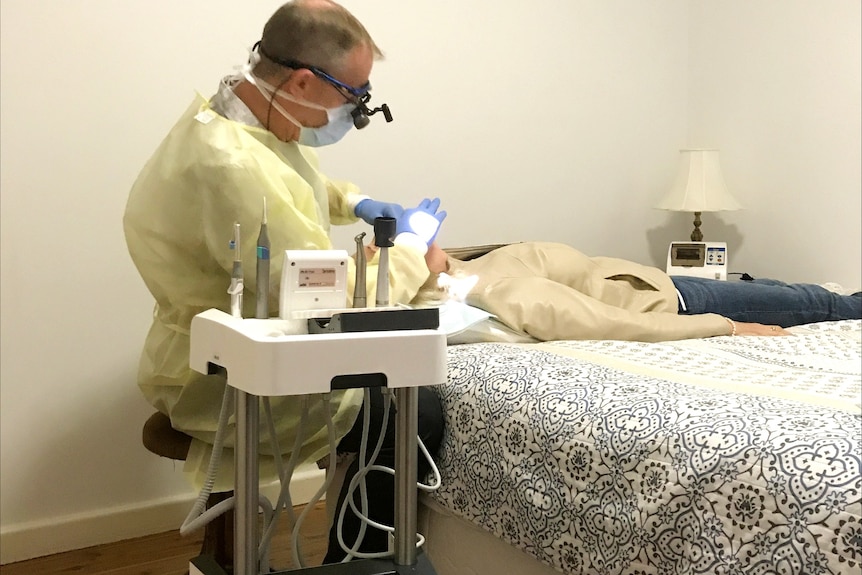 Man in yellow scrubs uses dental equipment on a woman lying on a bed